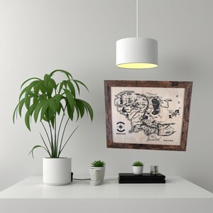 May include: A framed map of Middle-earth from The Lord of the Rings, with a brown wooden frame and a white background. The map shows the major locations of Middle-earth, including the Shire, Mordor, and Gondor. The map is surrounded by a white wall and a hanging light fixture.