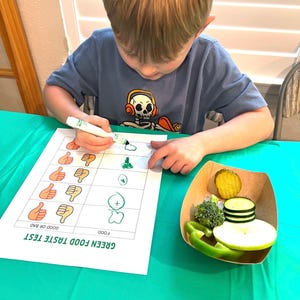 May include: A child is completing a green food taste test. The child is using a green marker to rate the food on a chart with thumbs up and thumbs down. The chart says "Green Food Taste Test" and has the words "Food" and "Good or Bad". The child is rating a plate of green food, including broccoli, cucumber, apple, and green pepper.