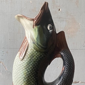 May include: A ceramic fish-shaped jug with a detailed scale pattern and fins. The jug has a dark brown head and tail, with a green and brown body. The handle is formed by the fish's tail, and the base is yellow and green.