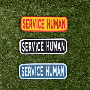 Op de afbeelding: Drie geborduurde patches met de tekst "SERVICE HUMAN" in witte letters. De patches zijn geel, zwart en blauw.