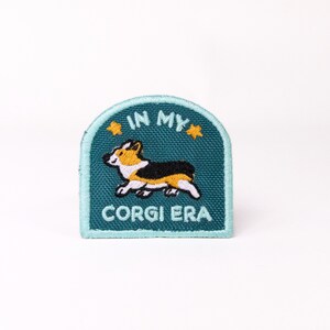 May include: A teal blue embroidered patch with a cartoon corgi running and the text "In My Corgi Era" with two yellow stars.