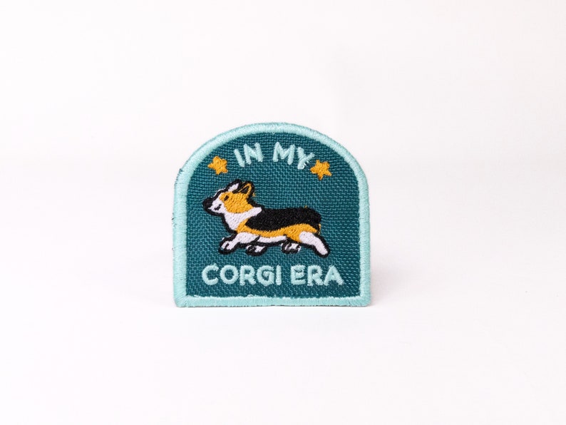 May include: A teal blue embroidered patch with a cartoon corgi running and the text "In My Corgi Era" with two yellow stars.