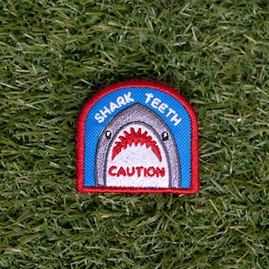 May include: Embroidered patch with a shark design. The patch features a shark's open mouth with sharp teeth, the text "SHARK TEETH" above, and "CAUTION" below. The patch has a blue, red, and white color scheme.
