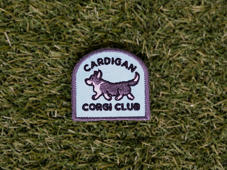May include: A light blue embroidered patch with a black and white cartoon Cardigan Welsh Corgi walking. The patch says "Cardigan Corgi Club" in black embroidery.