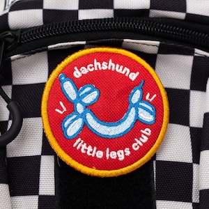 Könnte beinhalten: Nahaufnahme eines schwarz-weiß karierten Rucksacks mit einem runden Aufnäher. Der Aufnäher ist rot mit gelbem Rand und zeigt ein blaues Ballonhund-Design und die Worte "dachshund little legs club". Ein schwarzer Reißverschluss ist sichtbar.
