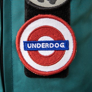 May include: A red, white, and blue embroidered patch with the word "UNDERDOG" in a blue banner inside a red circle. The patch is sewn onto a teal fabric background.