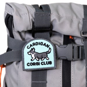 May include: A gray backpack with a black and orange zipper and a blue and white embroidered patch that says "CARDIGAN CORGI CLUB" with a cartoon corgi dog on it.