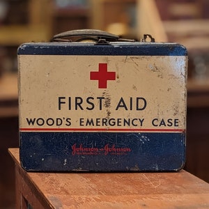 May include: Vintage metal first aid kit with a red cross and the text "FIRST AID WOOD'S EMERGENCY CASE" and the brand name "Johnson & Johnson".