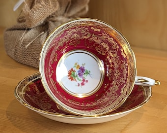 May include: A red teacup and saucer with gold trim and a floral design. The teacup and saucer are on a wooden surface.
