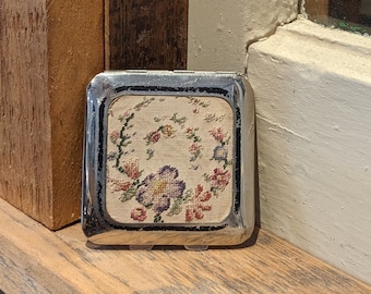 Puede incluir: Un pequeño estuche compacto con marco plateado y un diseño floral en el interior. El diseño presenta una variedad de flores en tonos de rosa, morado y rojo.
