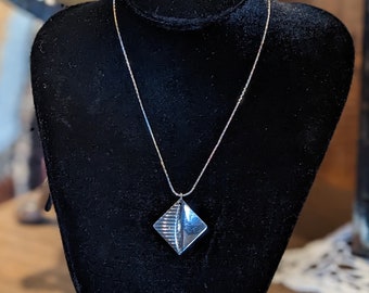 Puede incluir: Un collar de plata con un colgante geométrico. El colgante tiene forma de diamante con una superficie texturizada.