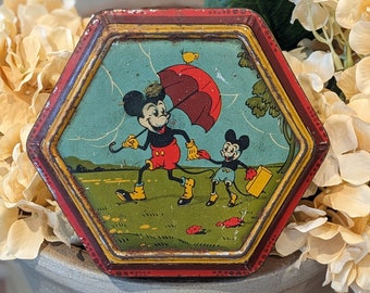 May include: Vintage hexagonal tin lunchbox featuring Mickey Mouse and a smaller Mickey Mouse holding an umbrella. The tin is red, yellow, and blue with a green background.