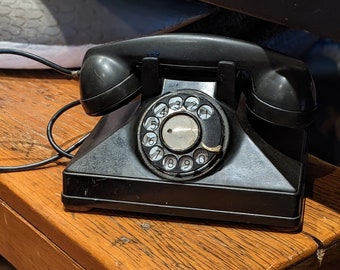 Könnte beinhalten: Ein schwarzes Wählscheibentelefon mit einem schwarzen Kabel auf einem Holztisch.