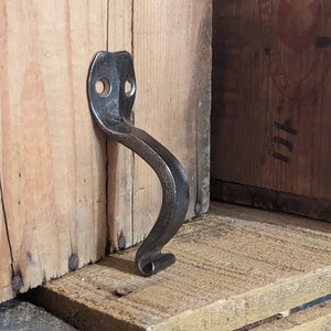 May include: A black metal handle with a curved design. The handle is attached to a wooden surface.