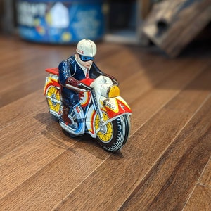 以下が含まれることがあります： 警察官のフィギュアが乗った、ビンテージのブリキのおもちゃのオートバイ。オートバイは赤、白、青で、側面に「PATROL」と印刷されています。警官は青い制服を着て、白いヘルメットと黒いマスクをしています。