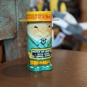 May include: A vintage tin toy bank shaped like a man wearing a yellow hat and green suit. The bank has a sign that reads "Watch Me Grow Tall With Coins Large & Small".