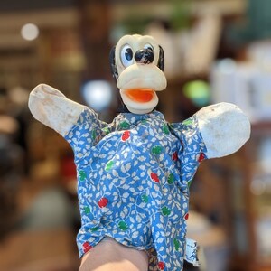 May include: A vintage hand puppet of a cartoon dog character. The puppet has a black and white head, an orange mouth, and a blue floral patterned shirt. The arms are made of off-white fabric.