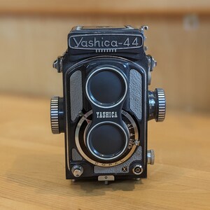 May include: A black Yashika-44 camera with a silver lens and a silver and black body. The camera has a silver dial with the word "YASHICA" printed on it.