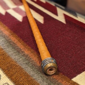 May include: A long, brown wooden pipe with a silver metal band and a wooden mouthpiece. The pipe is lying on a colorful striped blanket.