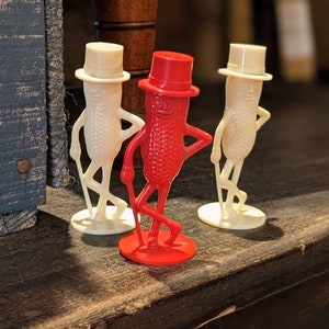 Peut inclure: Trois salières et poivrières en forme d'arachide. Deux sont blanches avec des chapeaux noirs et l'autre est rouge avec un chapeau noir.