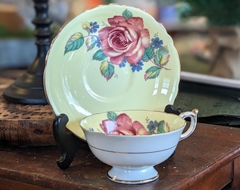 May include: A vintage teacup and saucer set with a floral design. The saucer is pale yellow with a pink rose and green leaves. The teacup is white with a pink rose and green leaves.