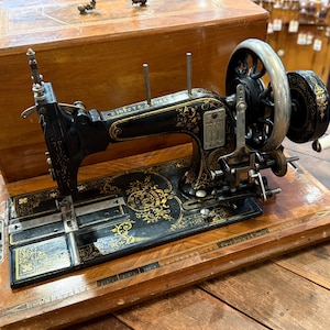 May include: A vintage black cast iron sewing machine with intricate gold floral decals and a silver hand crank wheel. The machine is mounted on a rectangular wooden base and includes a matching wooden cover with a metal handle on top.