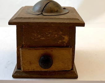 Op de afbeelding: Een vintage houten koffiemolen met een metalen handvat en een lade om koffiebonen op te bergen. De molen is bruin geverfd en heeft een verweerde, rustieke uitstraling.