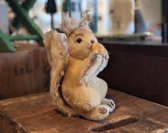 Könnte beinhalten: Ein Plüsch-Eichhörnchen mit einem flauschigen Schwanz, das auf einer Holzkiste sitzt. Das Eichhörnchen ist hellbraun und hat eine kleine, rosafarbene Nase.