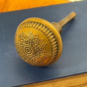 Puede incluir: Un pomo de puerta antiguo de latón con un diseño floral ornamentado. El pomo es redondo con un patrón detallado y un vástago rectangular. El pomo está sobre un libro azul con páginas doradas.