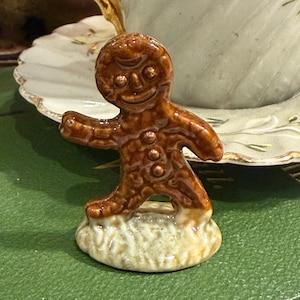 May include: A ceramic gingerbread man figurine with a smiling face, brown icing, and three button details. The figurine stands on a white base, set against a green background. A teacup is visible in the background.