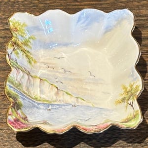 May include: A white porcelain dish with a scalloped edge and a painted scene of a coastal landscape with cliffs, water, and birds. The dish has a gold rim.