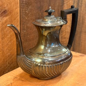 Pode incluir: Bule antigo prateado com uma pega preta. O bule tem um corpo bulboso com detalhes verticais canelados e um bico curvo. A tampa é encimada por um pequeno botão decorativo. O bule está exposto numa superfície de madeira.
