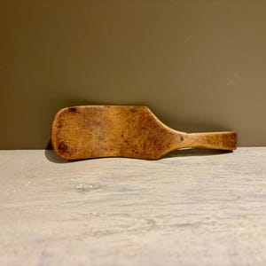 Könnte beinhalten: Ein Holzspatel mit einem breiten, rechteckigen Kopf und einem Griff. Der Spatel besteht aus hellbraunem Holz mit sichtbarer Maserung und einem leicht abgenutzten Aussehen. Der Griff ist abgerundet und verjüngt sich zum Ende hin.