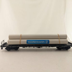 May include: A black model train car with two white pipes secured with rubber bands. The car has a blue label that reads "MANNESMANN" with a white logo.