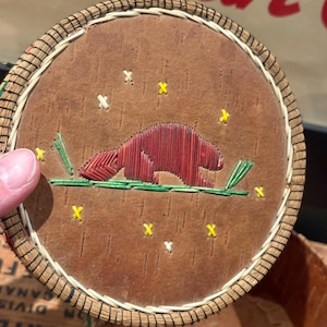 May include: Round, handcrafted coaster with a brown wooden base and a woven rim. The coaster features an embroidered design of a red beaver on a green branch, with yellow star accents. A unique, rustic home decor item.