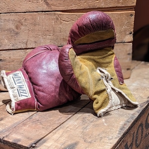 Puede incluir: Un par de guantes de boxeo vintage, uno rojo y otro amarillo, con el nombre de la marca "Winwell" impreso en el guante rojo. Los guantes est&aacute;n sobre una caja de madera.