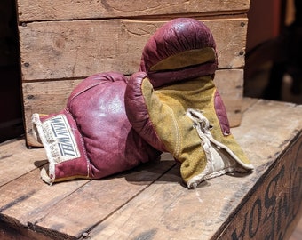 May include: A pair of vintage boxing gloves, one red and one yellow, with the brand name "Winwell" printed on the red glove. The gloves are sitting on top of a wooden crate.