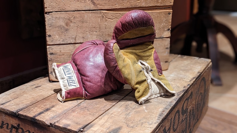 Puede incluir: Un par de guantes de boxeo vintage, uno rojo y otro amarillo, con el nombre de la marca "Winwell" impreso en el guante rojo. Los guantes est&aacute;n sobre una caja de madera.