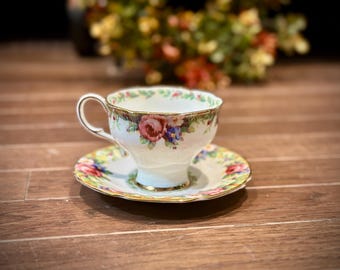 May include: A white teacup and saucer with a floral pattern in shades of pink, green, and blue. The cup has a gold rim and the saucer has a gold trim.