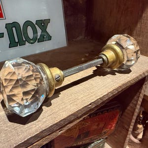 Può includere: Un pomello per porta in cristallo trasparente e sfaccettato con elementi in ottone. Il pomello è fissato a un'asta filettata e poggia su una mensola di legno. Lo sfondo include un'insegna con la scritta "R-NOX".