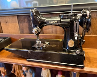 May include: A vintage black Singer sewing machine with gold accents. The machine is displayed on a wooden shelf. The Singer logo is visible on the machine's body. The machine has a hand wheel and a needle bar.