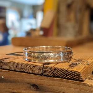 May include: A silver bangle bracelet with an engraved floral design and a chain attached to the side.