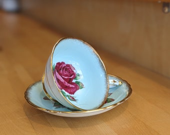 May include: A light blue teacup and saucer with a gold rim. The teacup features a single red rose with green leaves printed on it.