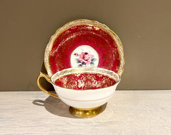 May include: A vintage teacup and saucer set with a red and gold floral pattern. The cup and saucer are decorated with a delicate floral design and a gold trim.