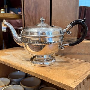 May include: A silver-plated teapot featuring a polished metallic finish, a curved spout, a circular lid with a small finial, and a black C-shaped handle. The body is decorated with a horizontal band of embossed detailing and rests on a pedestal base.