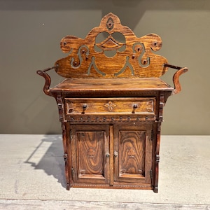 Puede incluir: Un pequeño gabinete de madera tallada con un cajón y dos puertas. El gabinete está hecho de madera oscura con un rico patrón de veta. La parte superior del gabinete presenta un diseño decorativo calado.