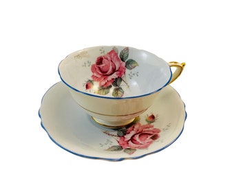 May include: A vintage teacup and saucer set. The cup and saucer are white with a pink rose floral design and gold trim. The rim of the cup and saucer are trimmed with a blue border. The handle of the cup is gold.