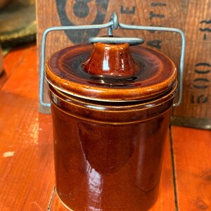 Peut inclure: Petit pot en céramique marron avec couvercle et anse métallique. Le pot a une finition brillante et une forme cylindrique. Le couvercle est légèrement bombé et s'adapte parfaitement. L'anse est en métal argenté.