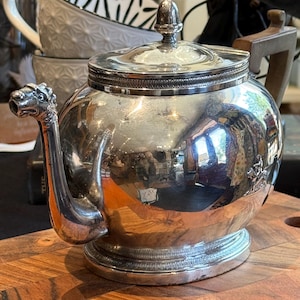 Peut inclure: Une théière argentée polie avec un corps arrondi, un couvercle surmonté d'un petit bouton et un bec verseur en forme de tête d'animal. La base et le bord du couvercle présentent un motif texturé, avec une petite figurine en relief sur le côté.