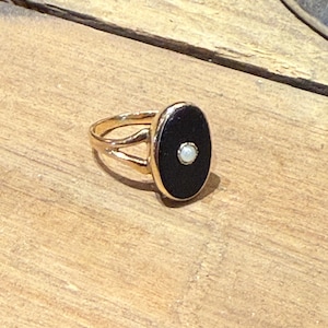 Peut inclure: Une bague dor&eacute;e avec une pierre noire ovale et une petite perle blanche ronde au centre. La bague a un design &agrave; anneau fendu. La bague est pos&eacute;e sur une surface en bois.